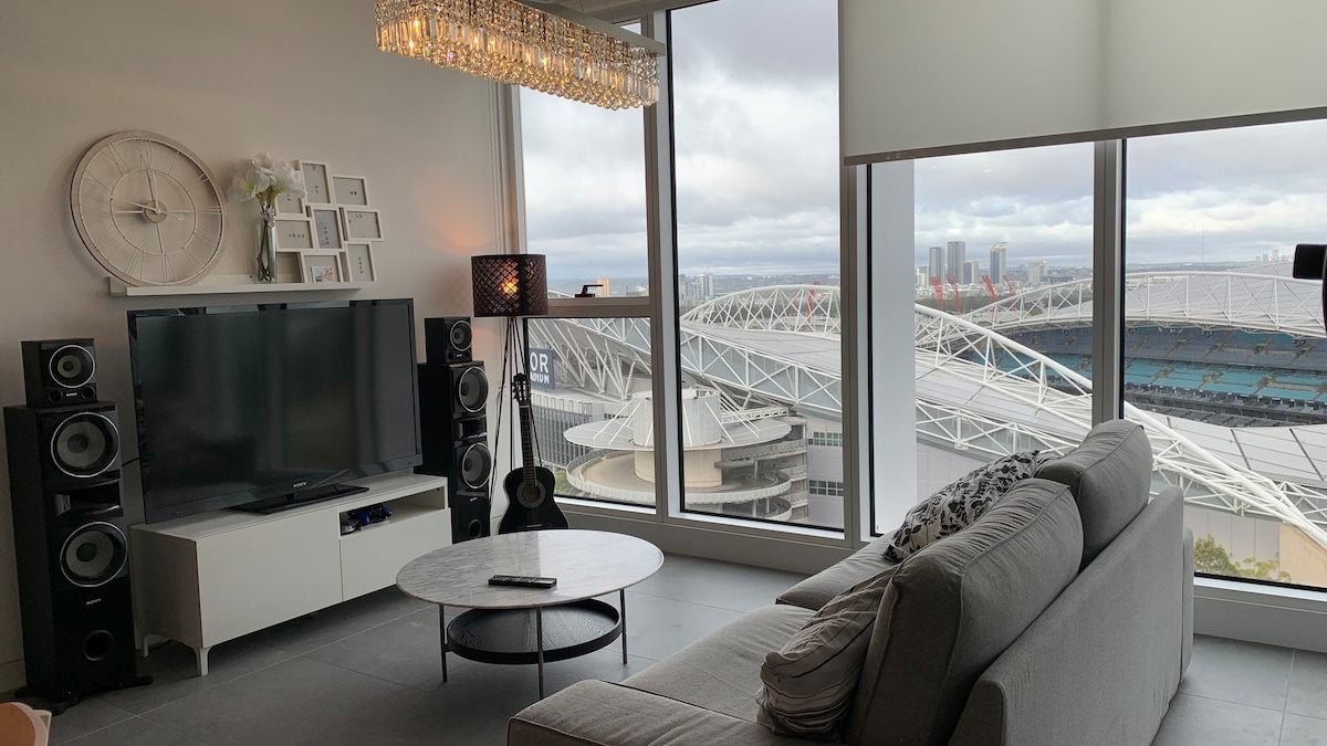 A stylish living area features a large flat-screen TV mounted on a white media console. A modern gray sofa is positioned facing the TV, complemented by a coffee table. Large windows offer a panoramic view of the ACCOR stadium and city skyline.