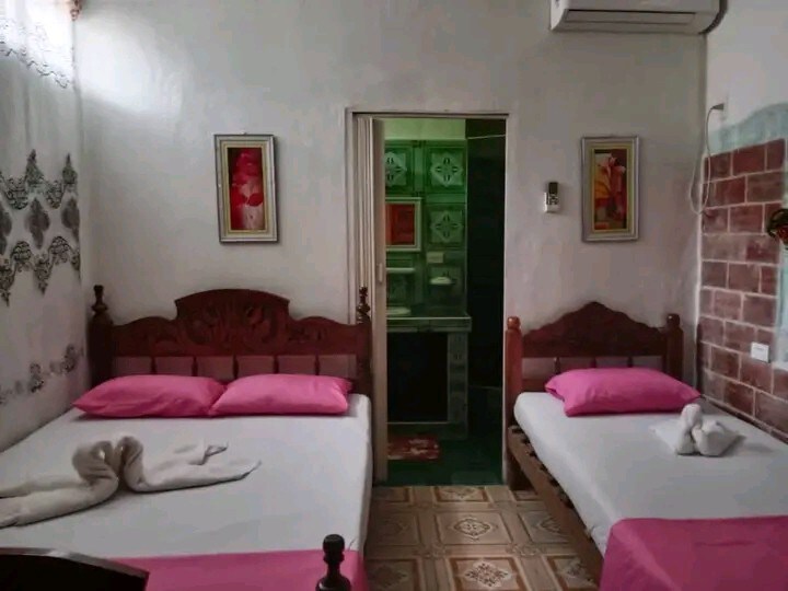 A comfortable room is presented with two twin beds, each adorned with pink pillows. Artwork is displayed on the walls, and a doorway leads to a bathroom area. Subtle patterned tiles cover the floor, contributing to the cozy ambiance.