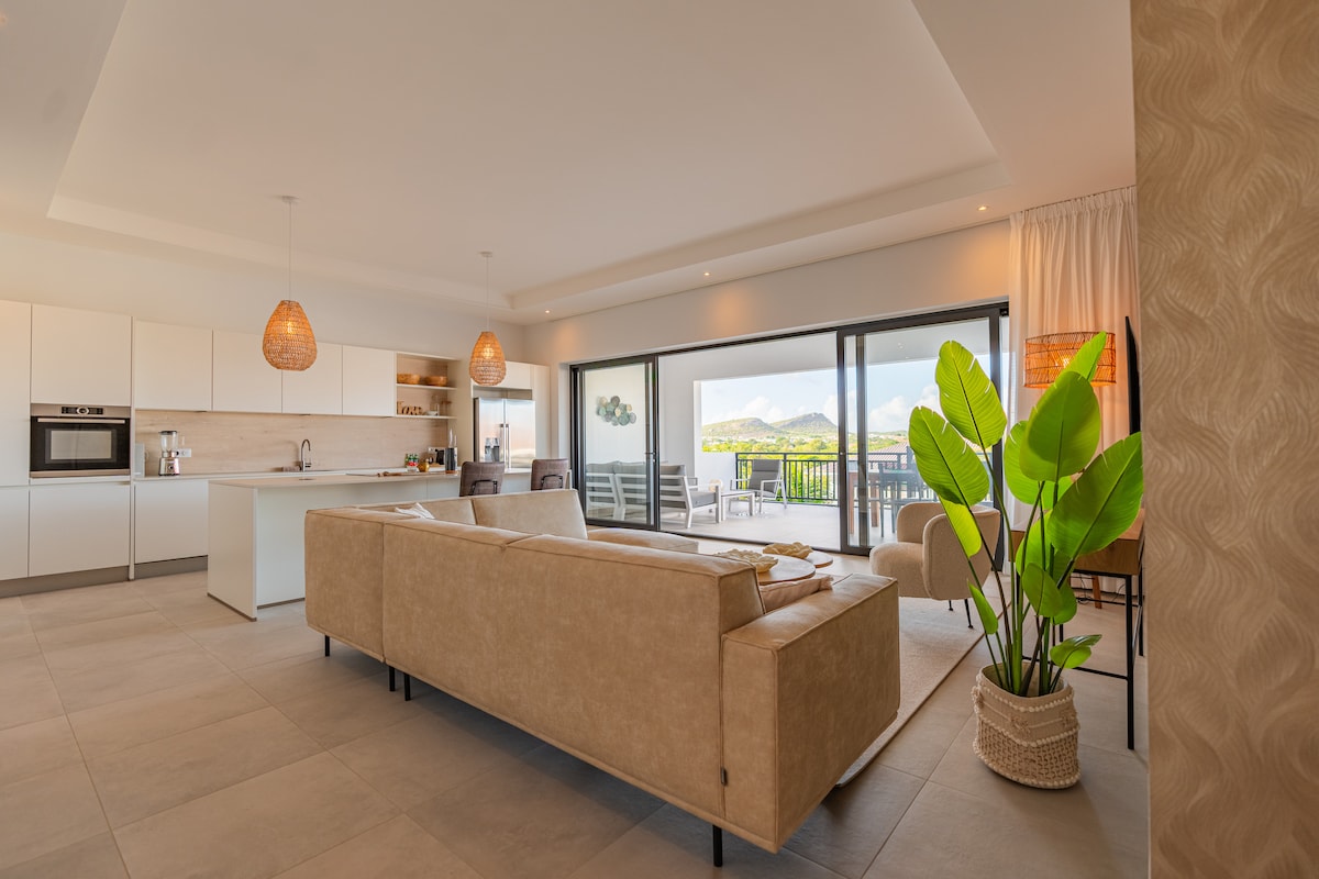 A spacious living area features a soft sofa and modern kitchen, with large glass doors offering natural light and access to a private balcony. Indoor plants add a touch of greenery, while the view of distant mountains is visible through the windows.