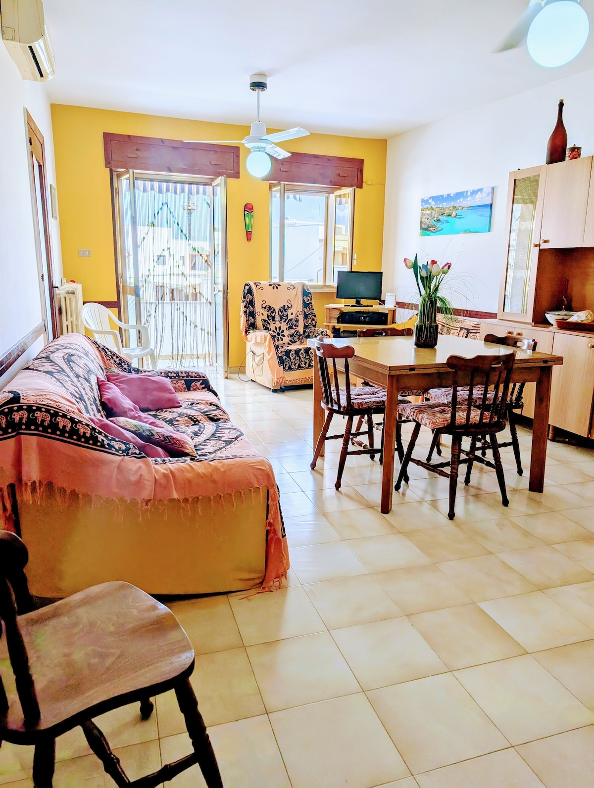 A living area with a bright yellow wall creates a warm ambiance. Two couches are positioned adjacent to a dining table surrounded by four chairs. Light from the balcony illuminates the space, and a small wall-mounted decor piece adds character.