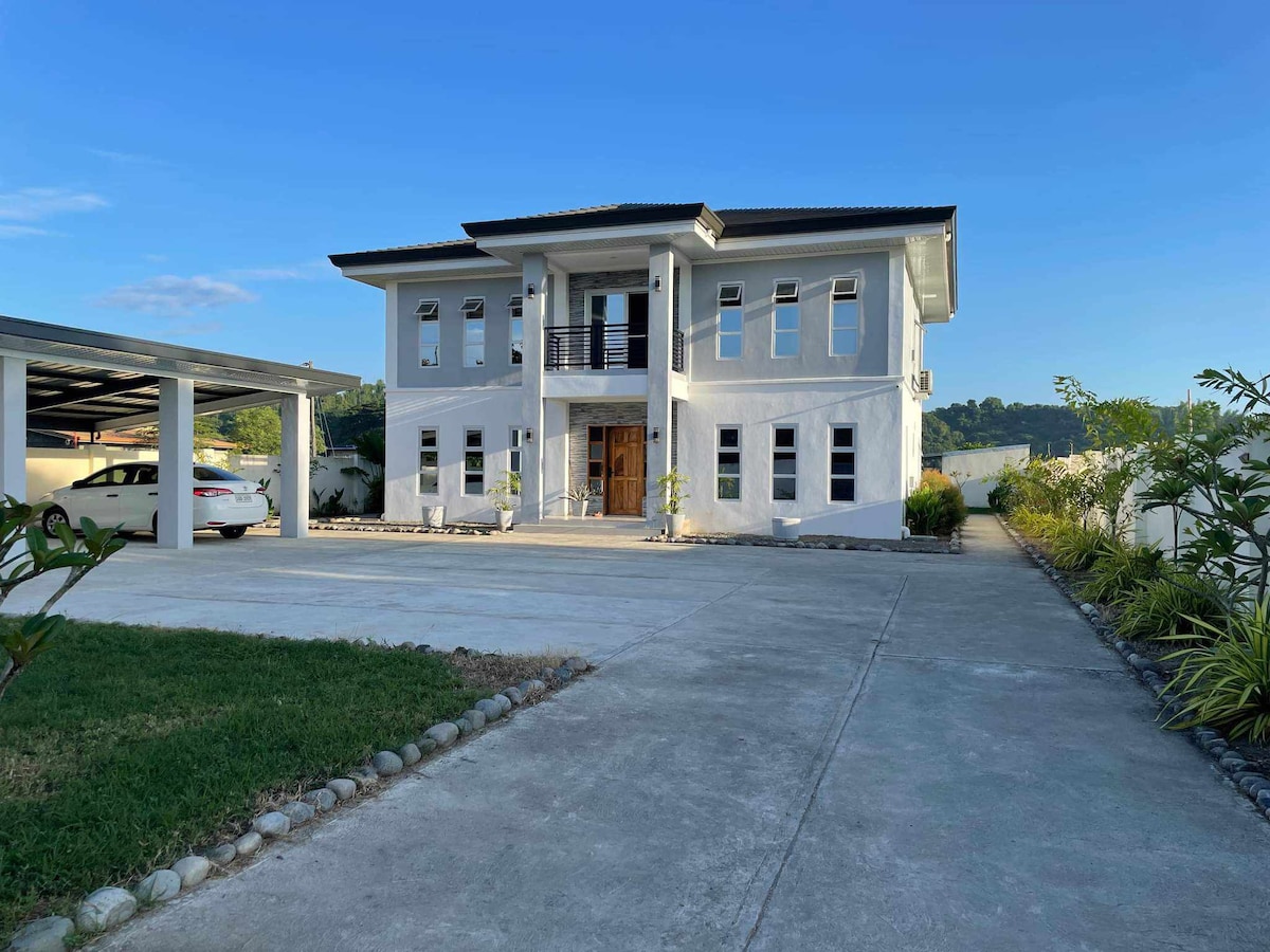 A modern two-story guest house is displayed with a spacious driveway leading to a welcoming entrance. The exterior features light-colored walls, tall windows, and a balcony on the upper level. Lush greenery and a paved pathway accentuate the surrounding area.