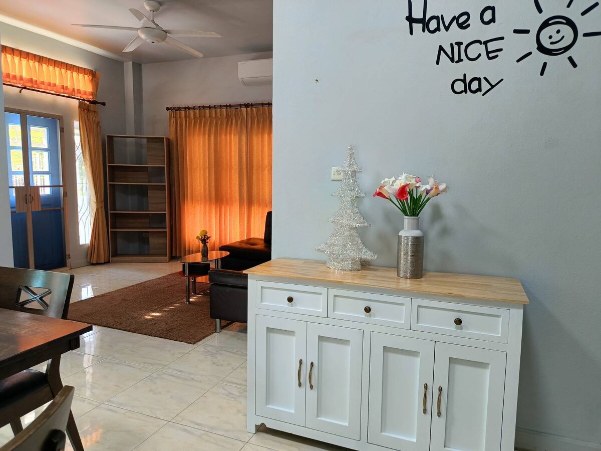 A light-filled living area is highlighted by large windows with flowing curtains. A white sideboard features a decorative arrangement of flowers and a small decorative tree. A cozy couch and a bookcase are positioned in the background, contributing to a welcoming environment.