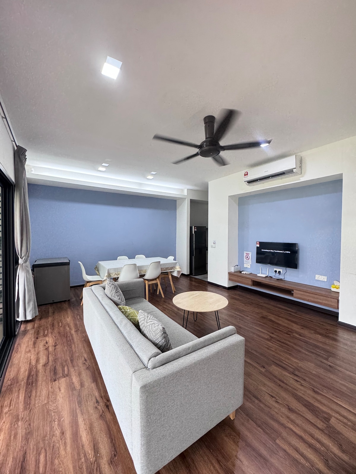 A spacious living area features a light gray sofa with decorative cushions, facing a flat-screen TV mounted on a wooden panel. A dining area with a white table and six chairs is visible in the background. Large windows allow natural light to enter.