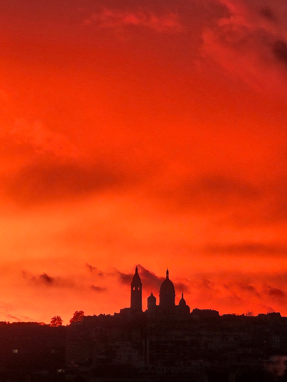 The silhouette of Paris’s iconic Sacré-Cœur Basilica stands against a vibrant orange sunset sky. The dramatic clouds create a striking contrast, highlighting the architectural details of the basilica's domes and towers.
