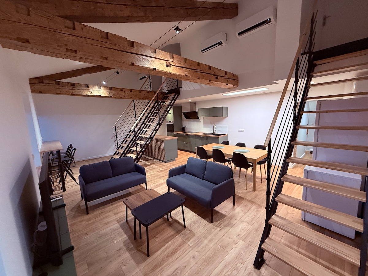 An open living area features two dark sofas facing a wooden coffee table. A dining table with chairs is positioned nearby, while modern kitchen appliances are visible in the background. Exposed wooden beams and stairs add architectural interest to the spacious design.