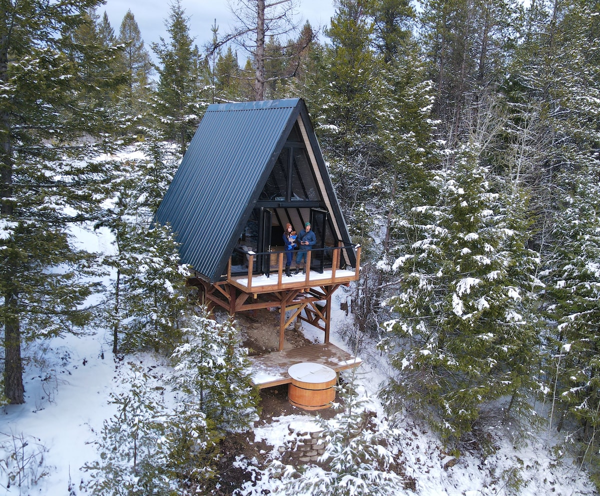 An A-frame cabin is nestled among snow-covered trees, elevated on stilts. A wooden deck extends from the front, featuring two chairs for outdoor seating. A hot tub can be seen in the foreground, surrounded by a stone patio and scattered snow.