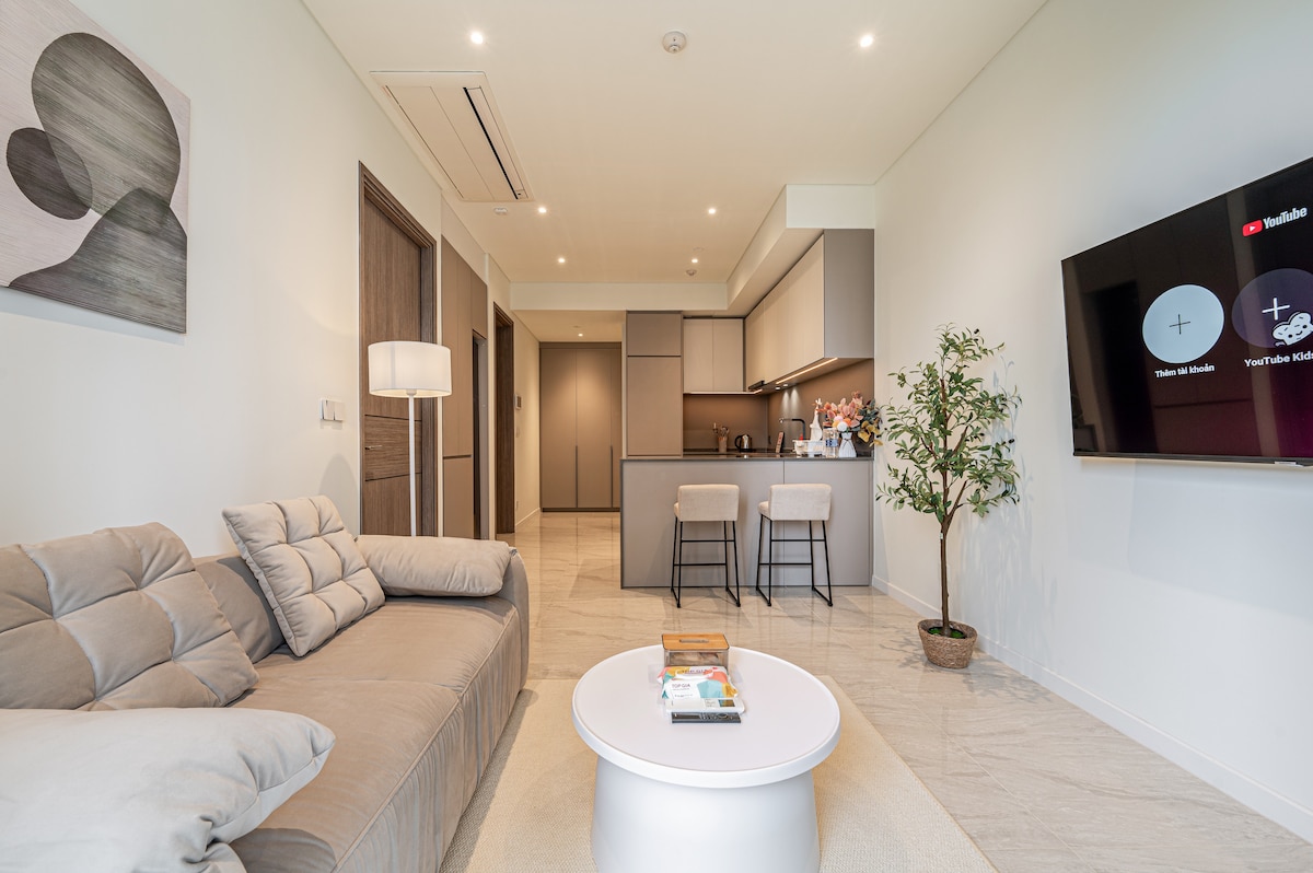 A modern living area features a comfortable sofa and a coffee table. The kitchen area is visible in the background, showcased with sleek cabinetry and bar stools. Soft lighting enhances the neutral color palette, while a potted plant adds a touch of greenery.