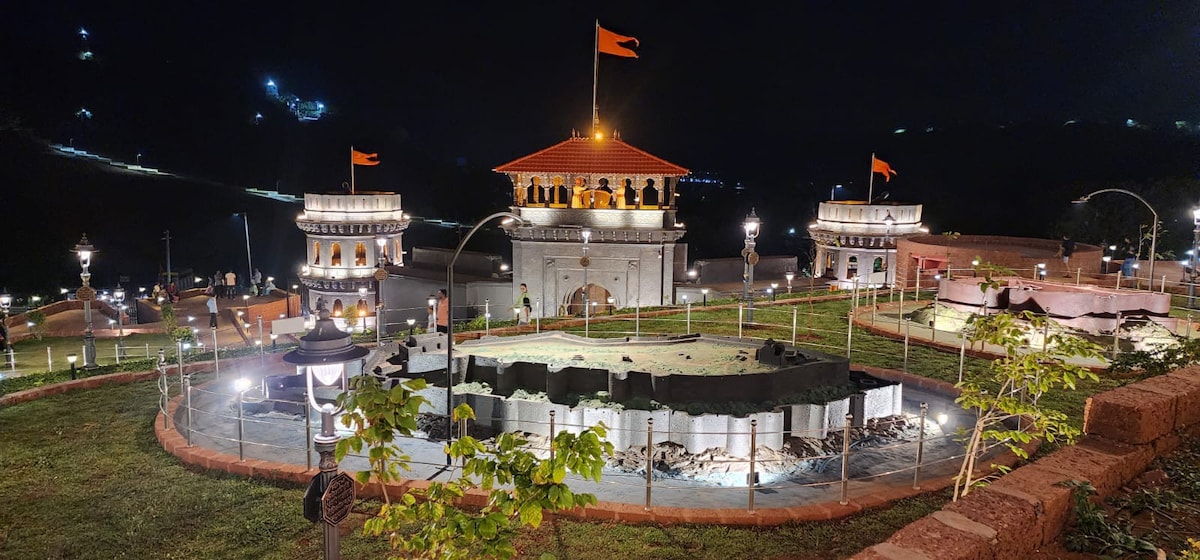 An illuminated fort structure is surrounded by lush greenery at night, with decorative lighting enhancing the architecture. The scene includes pathways and landscaped areas leading to the entrance, while vibrant flags flutter in the gentle breeze, adding a touch of color to the tranquil setting.