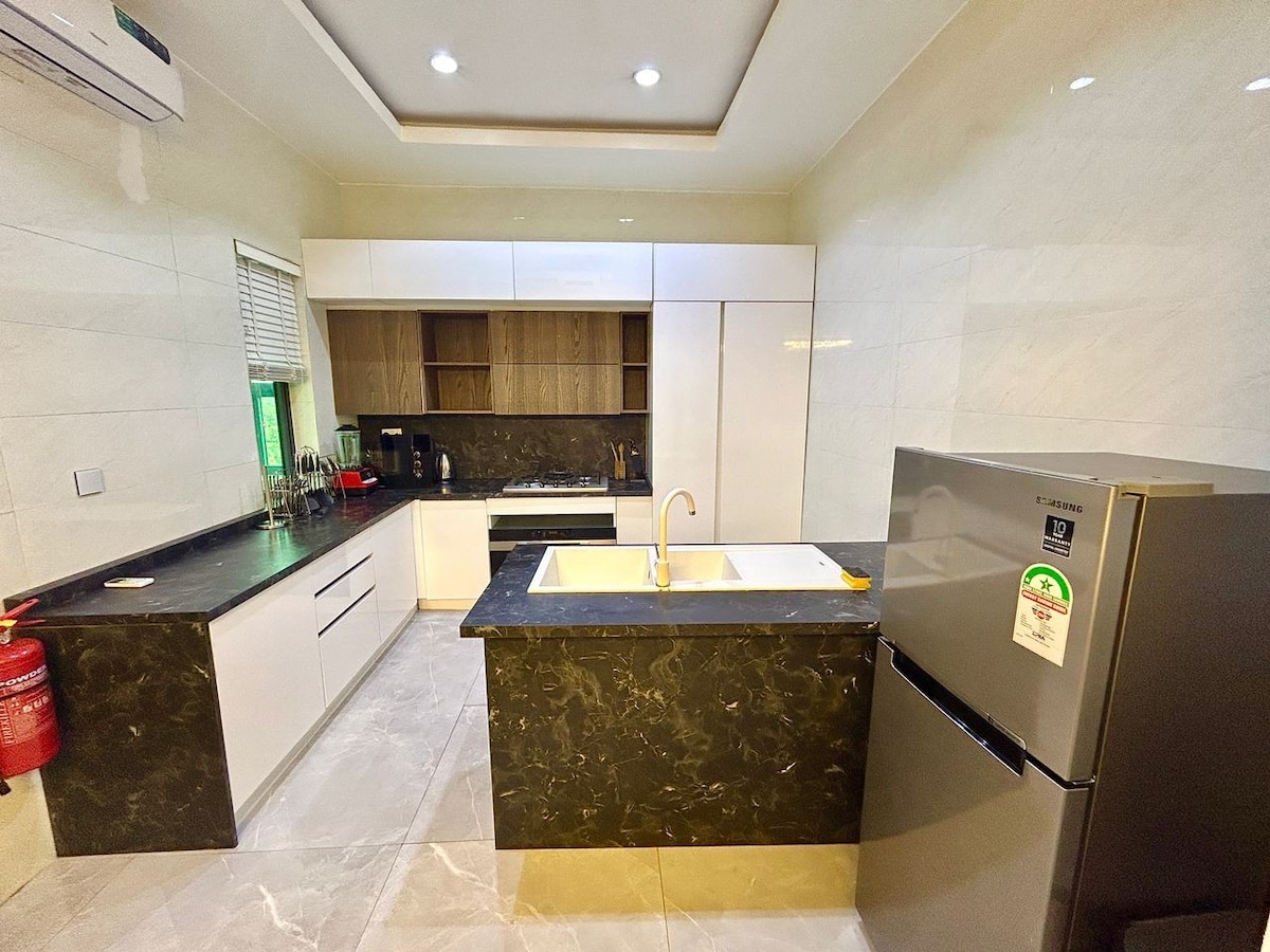 A modern kitchen is featured, showcasing sleek cabinetry and a dark countertop. Equipped with a refrigerator and a sink, the space offers ample storage and counter space. Natural light illuminates the area through a window, highlighting the clean, minimalist design.