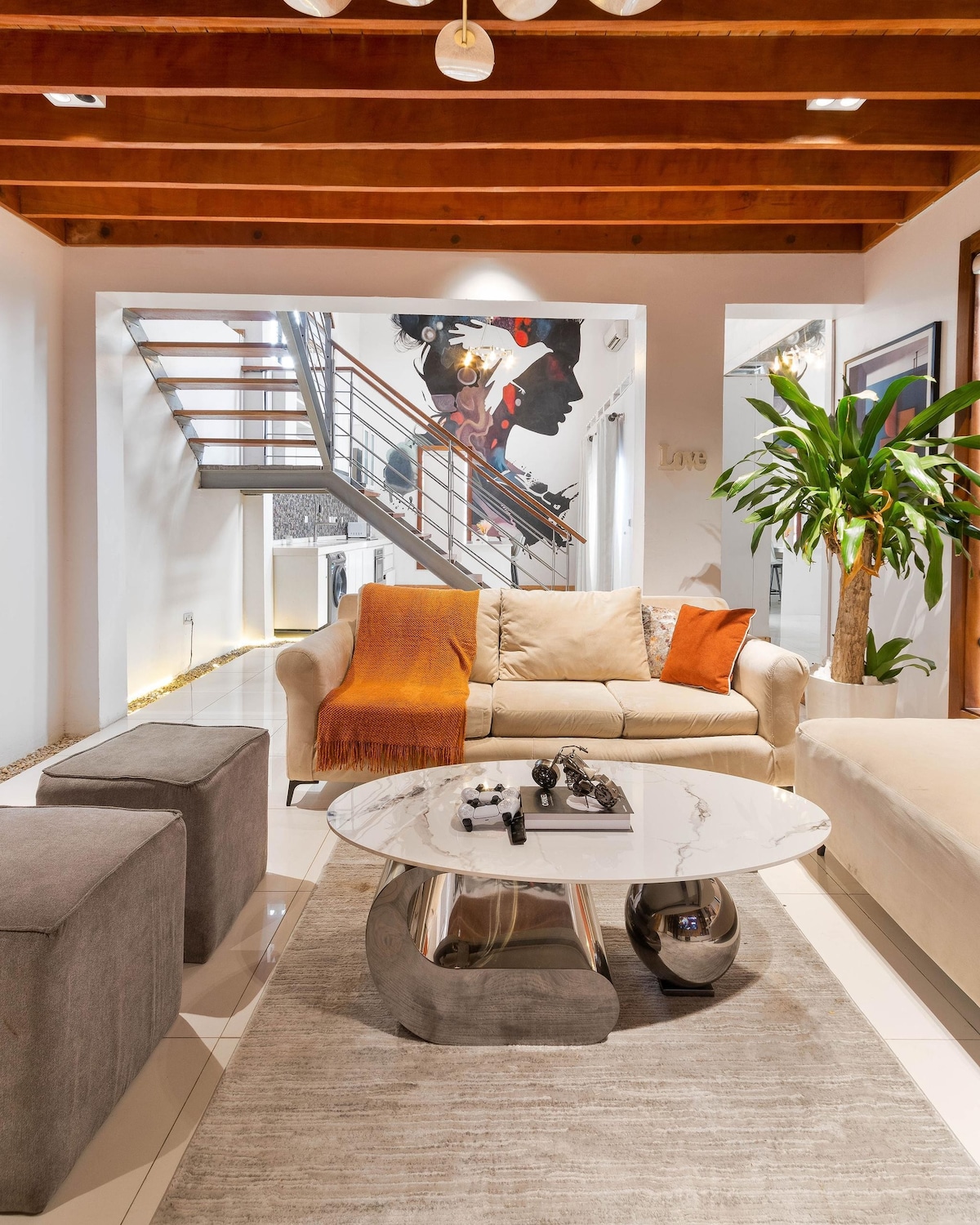 A spacious living area features a comfortable sofa draped with an orange throw, positioned next to a round marble coffee table. A decorative rug underfoot complements the grey ottomans. Artistic wall decor and a staircase with wooden accents add character to the room.