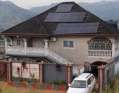 A large house features a distinctive roof adorned with solar panels. The exterior showcases a blend of brick and decorative wrought iron elements. A white vehicle is parked in front of the entrance, with landscaped greenery visible at the base of the property.
