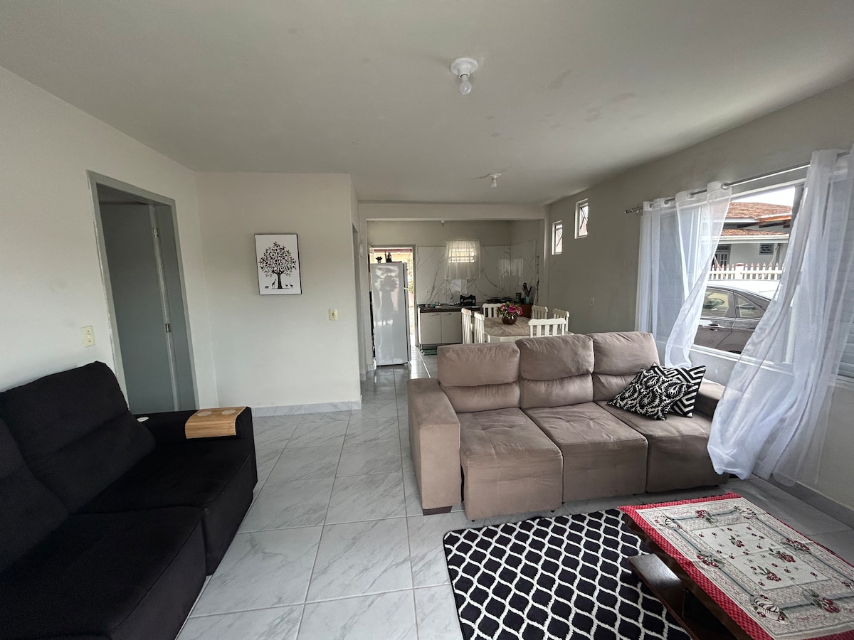 The living area features a spacious sofa and a dark armchair, arranged invitingly. A patterned area rug lays on the tiled floor. Natural light enters through large windows adorned with sheer curtains, creating a bright atmosphere. In the background, a kitchen area is visible.