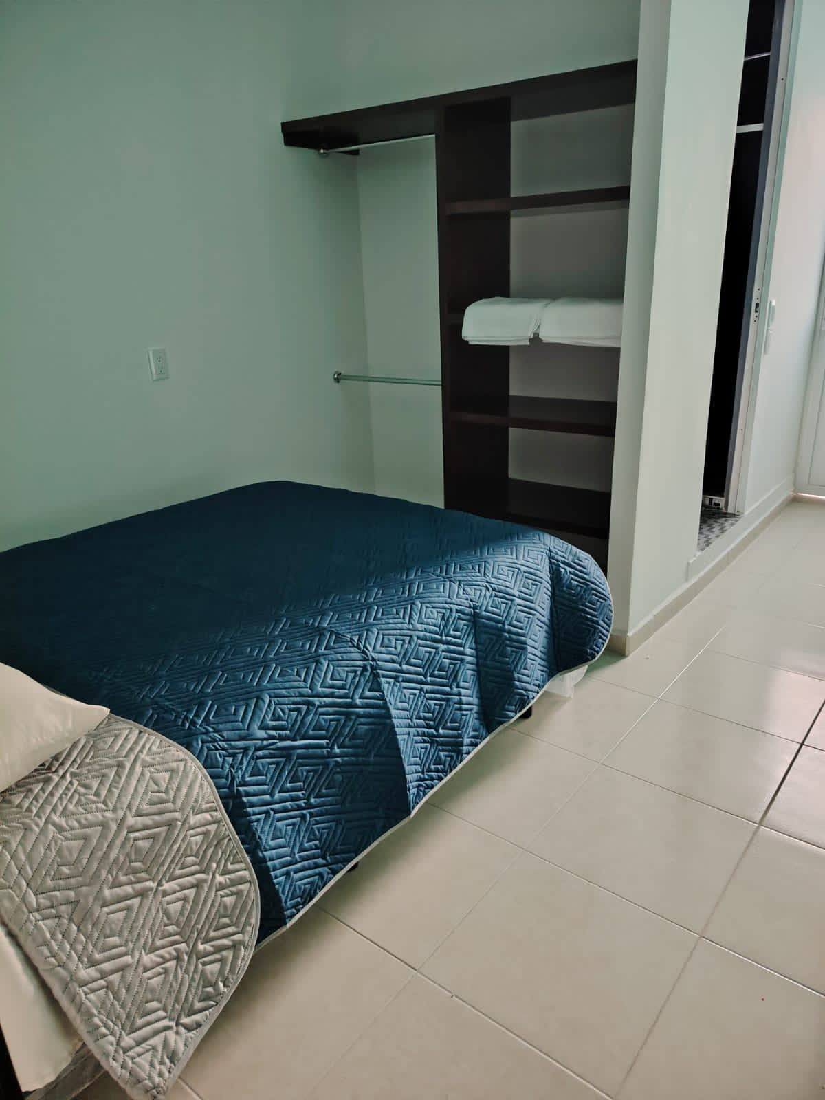 A well-lit bedroom features a spacious bed covered with a blue quilt. A closet with shelves is positioned against the wall, containing folded towels. Light-colored tiles reflect natural light coming from a nearby window.