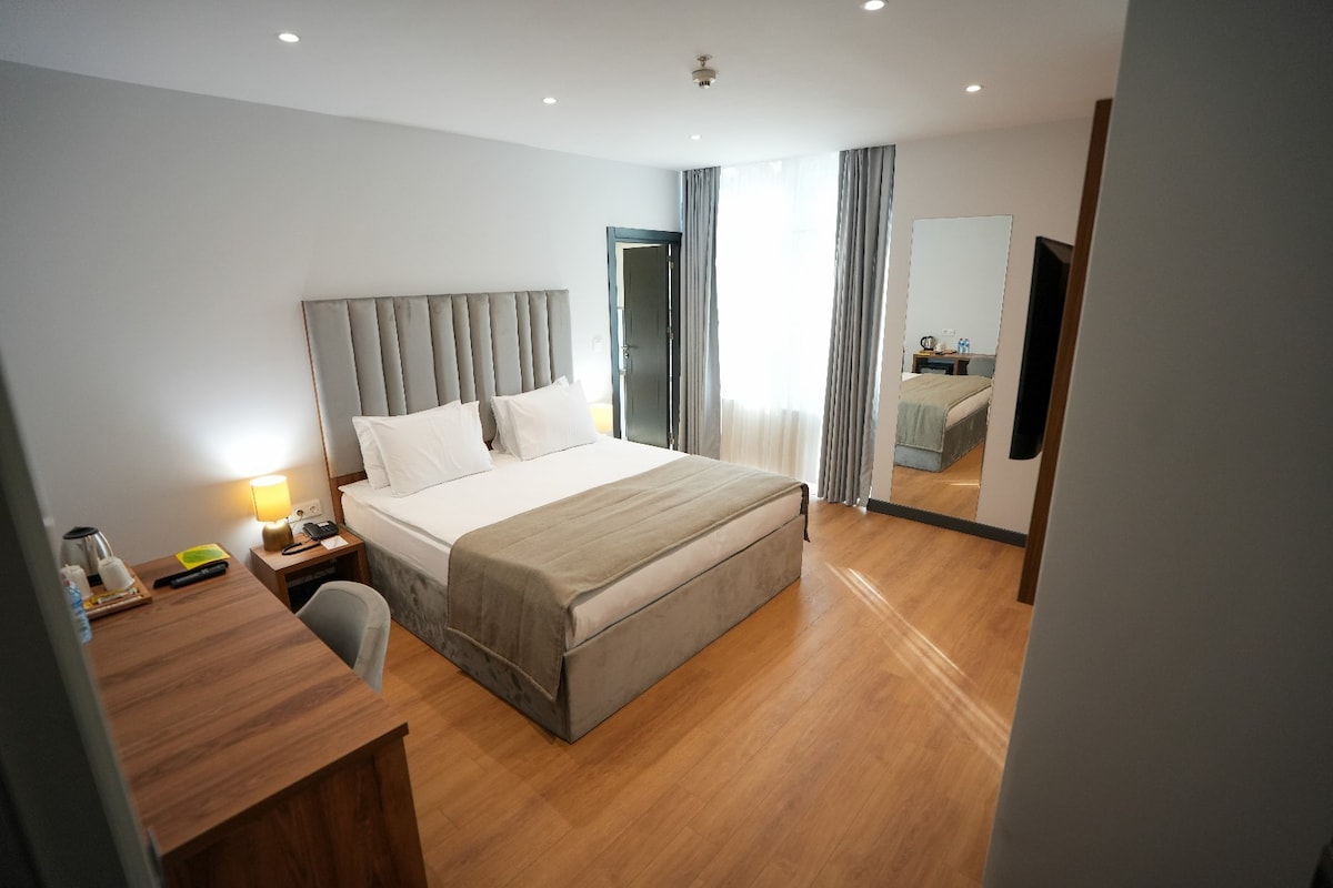 A comfortable guest room is showcased with a spacious bed, flanked by bedside lamps. Natural light filters through sheer curtains, illuminating the warm wooden flooring. A large mirror reflects the room's modern decor, which includes a desk and a kettle on a nearby table.