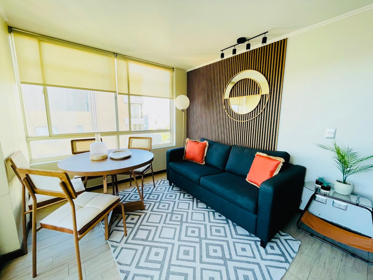 A modern living space features a dark sofa adorned with orange cushions. A round dining table is accompanied by four wooden chairs. An area rug with geometric patterns is positioned underneath the furniture. Natural light enters through large windows equipped with shades.