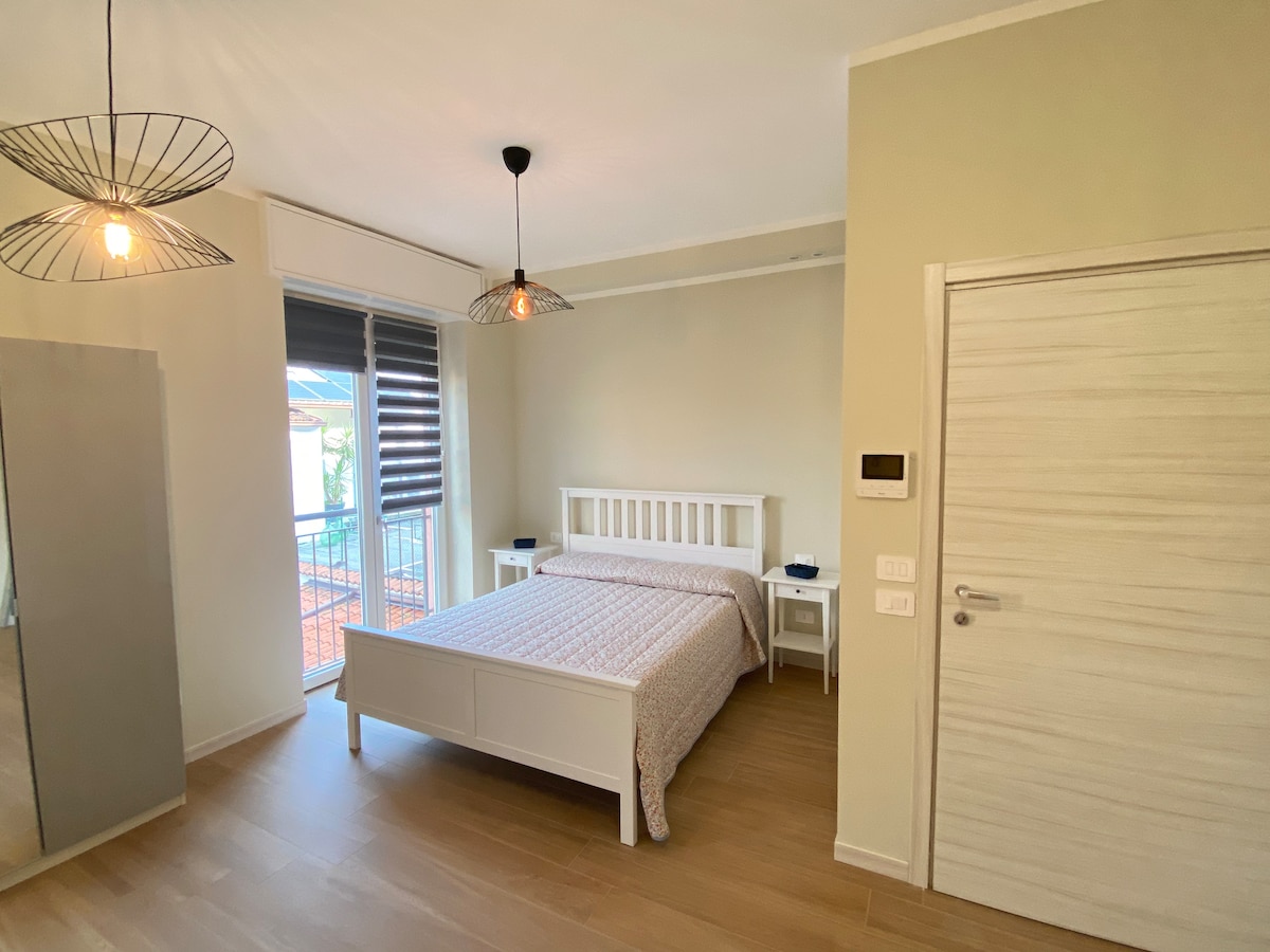 A bright sleeping area features a spacious bed with a light-colored patterned coverlet. Two pendant lights hang above, illuminating the room. A glass door with slatted blinds opens to a small balcony, while a full-length mirror stands against the wall opposite the bed.