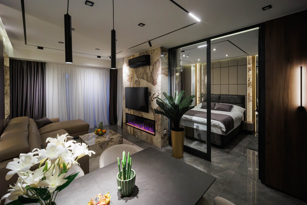 A modern living area features a plush, light-colored sofa and a dining table adorned with fresh flowers and greenery. A well-lit bedroom is partially visible through glass doors, showcasing a spacious bed. Soft lighting highlights the elegant design elements throughout the room.