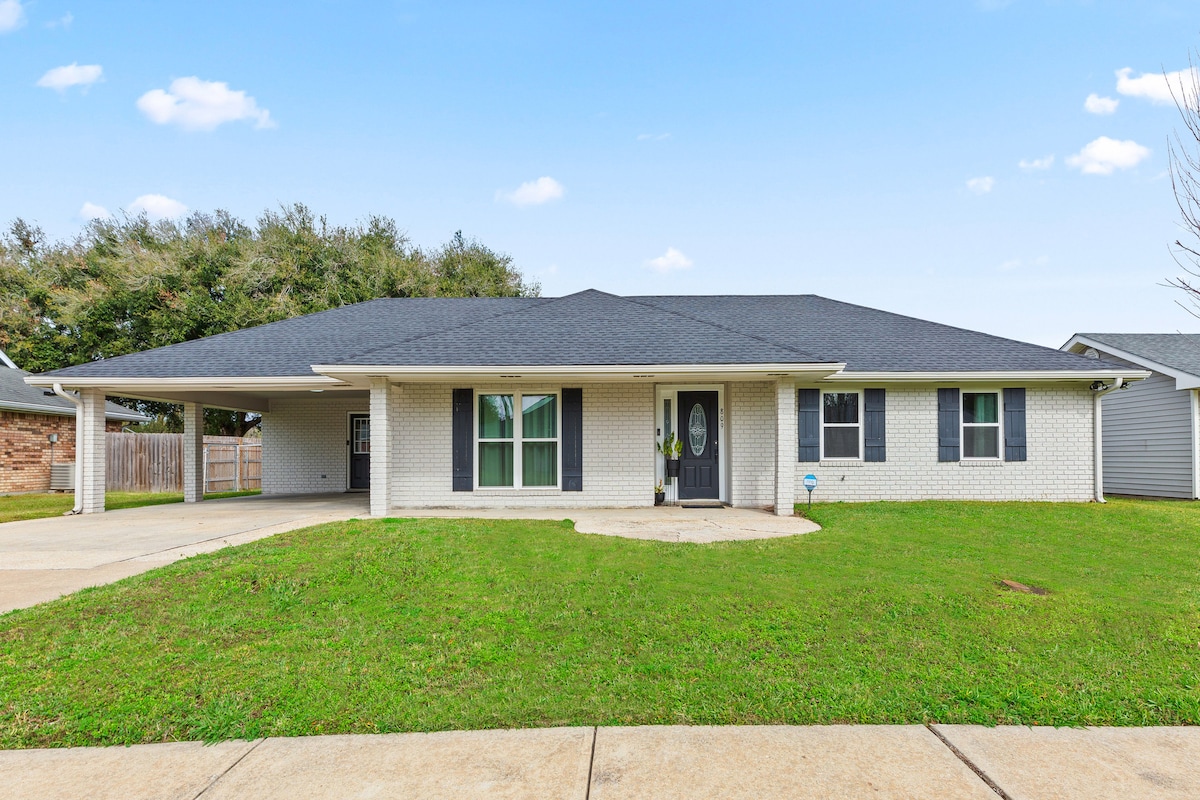 The exterior view of a ranch-style home features a covered porch with a circular entryway. Green grass surrounds the house, complemented by a concrete driveway. A large, shaded area provides space for outdoor activities, while trees frame the property in the background.