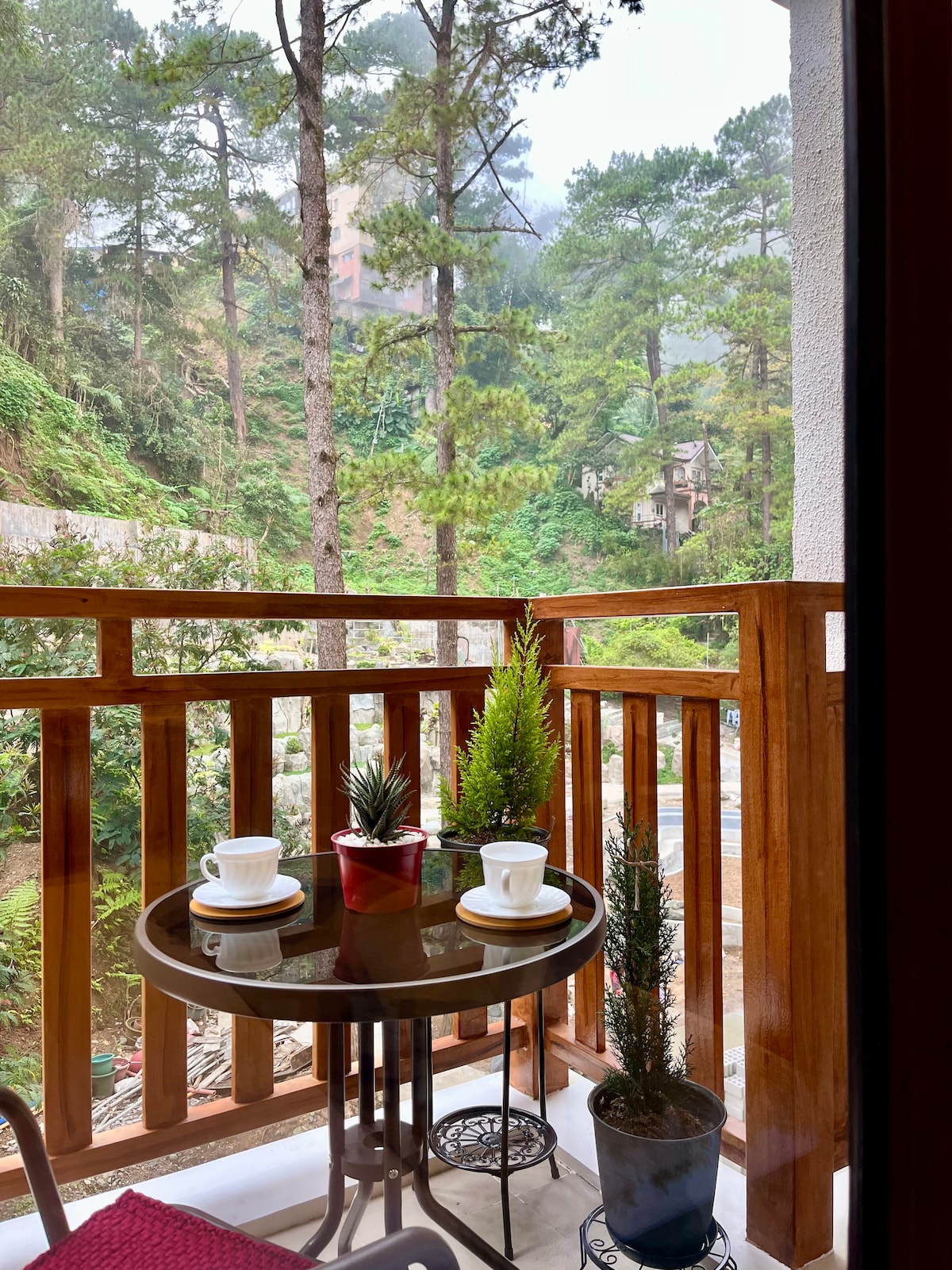 A small balcony features a round glass table accompanied by two cups and a small plant in a pot, creating an inviting space. Lush greenery and tall trees surround the area, enhancing the natural ambiance and providing a glimpse of the mountain landscape.