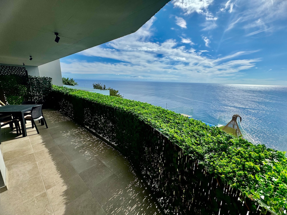 A spacious balcony overlooks the vast Atlantic Ocean, surrounded by lush greenery. Natural light brightens the area, highlighting outdoor seating arrangements. The serene view of the water and sky creates a peaceful atmosphere for relaxation.