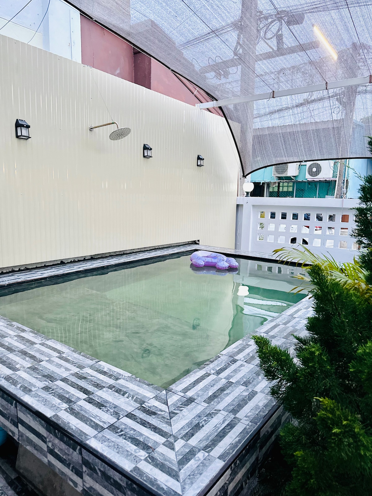 A private swimming pool is framed by smooth stone tiles, featuring a tranquil water surface. Adjustable shade structures provide sun protection. The pool area is enclosed by a white fence, creating a peaceful environment for relaxation and leisure activities.