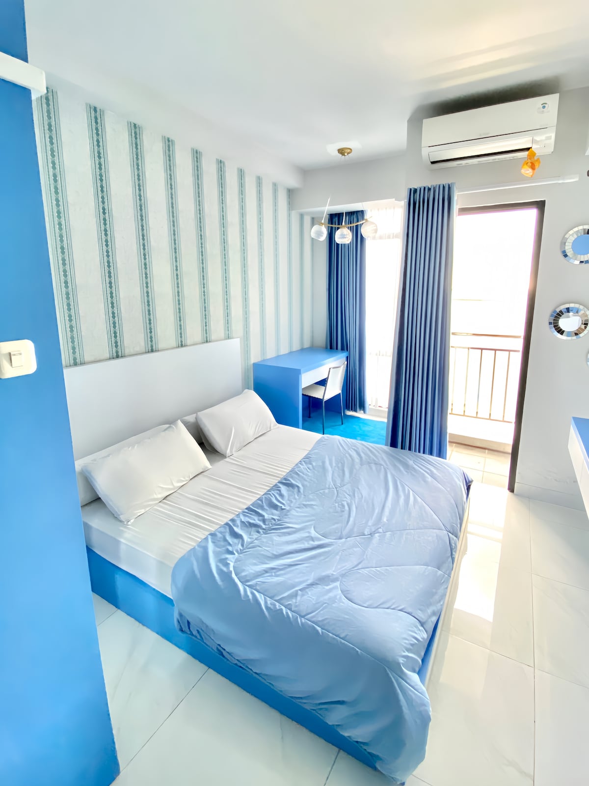 A bright sleeping area features light blue walls and soft natural light streaming through balcony doors. A spacious bed is dressed in a pale blue coverlet, complemented by a sleek work desk and a wall-mounted mirror, creating a serene and functional environment.