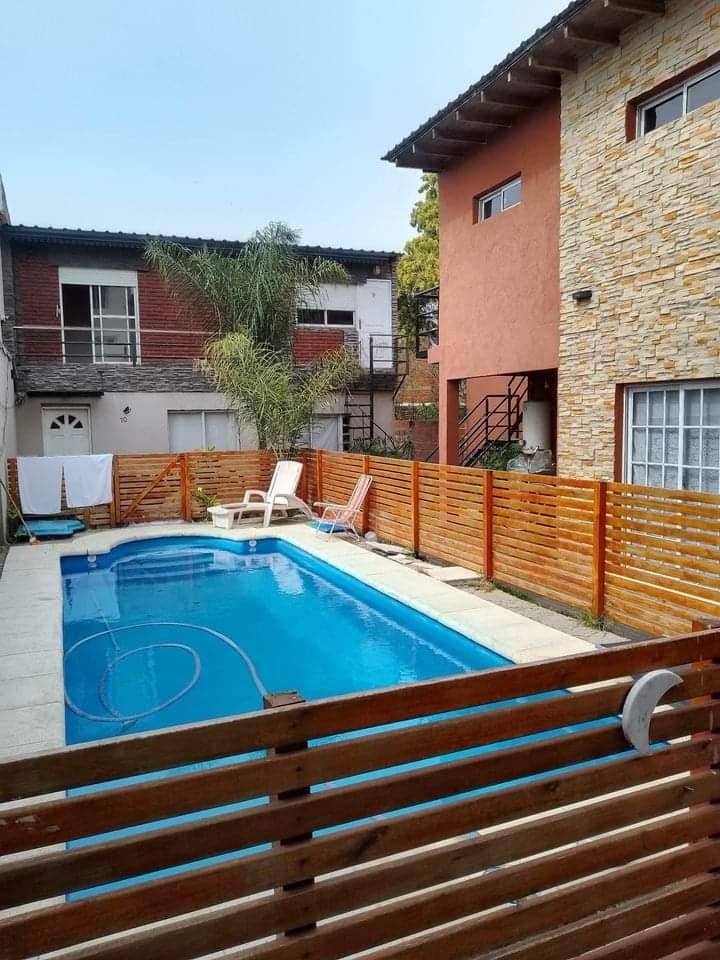 A private pool area is enclosed by wooden fencing, featuring a clear blue pool and two lounge chairs positioned on the side. The surrounding area shows a mix of buildings, with a staircase visible on one side and a pathway leading to the pool.