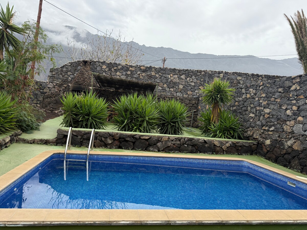 A serene outdoor area features a private swimming pool with clear blue water. Surrounding the pool, lush green plants and stone walls add a natural feel. The backdrop showcases distant mountains under a cloudy sky, enhancing the peaceful ambiance.