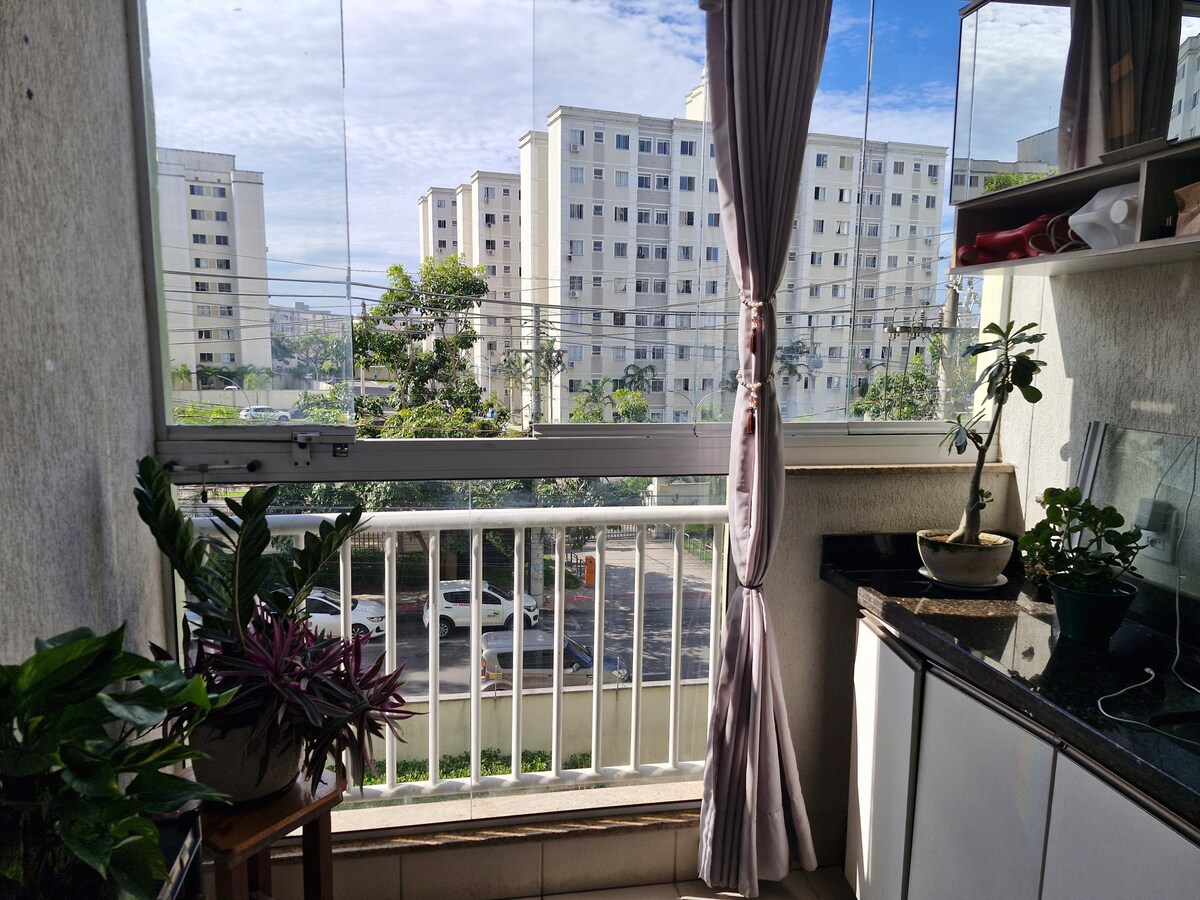 A kitchen area features a large window with glass panes that opens up to a balcony. Potted plants adorn the space, adding greenery. The counter is equipped with kitchen essentials while an outside view reveals nearby buildings and trees.