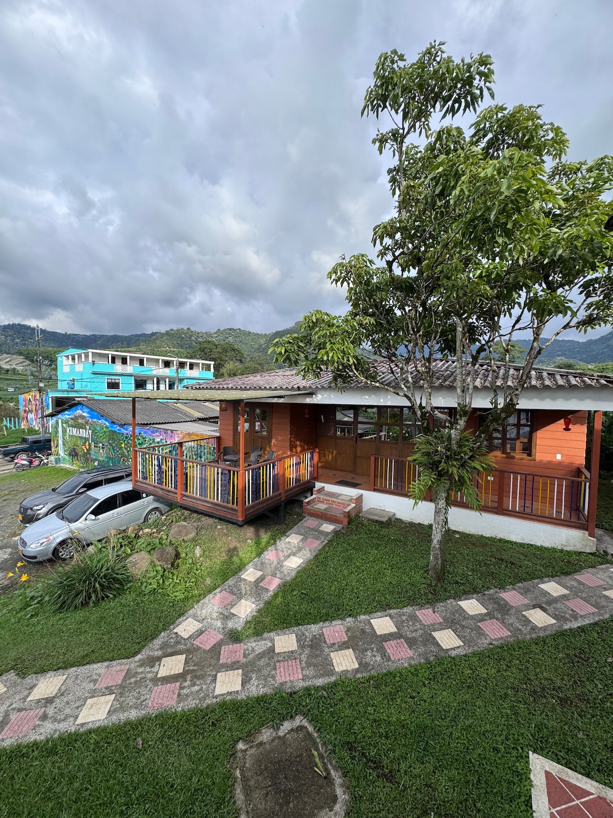A charming cabin is framed by a grassy area, featuring a pathway made of stone tiles leading to the entrance. Surrounding trees offer shade, while colorful buildings are visible in the background against a cloudy sky.