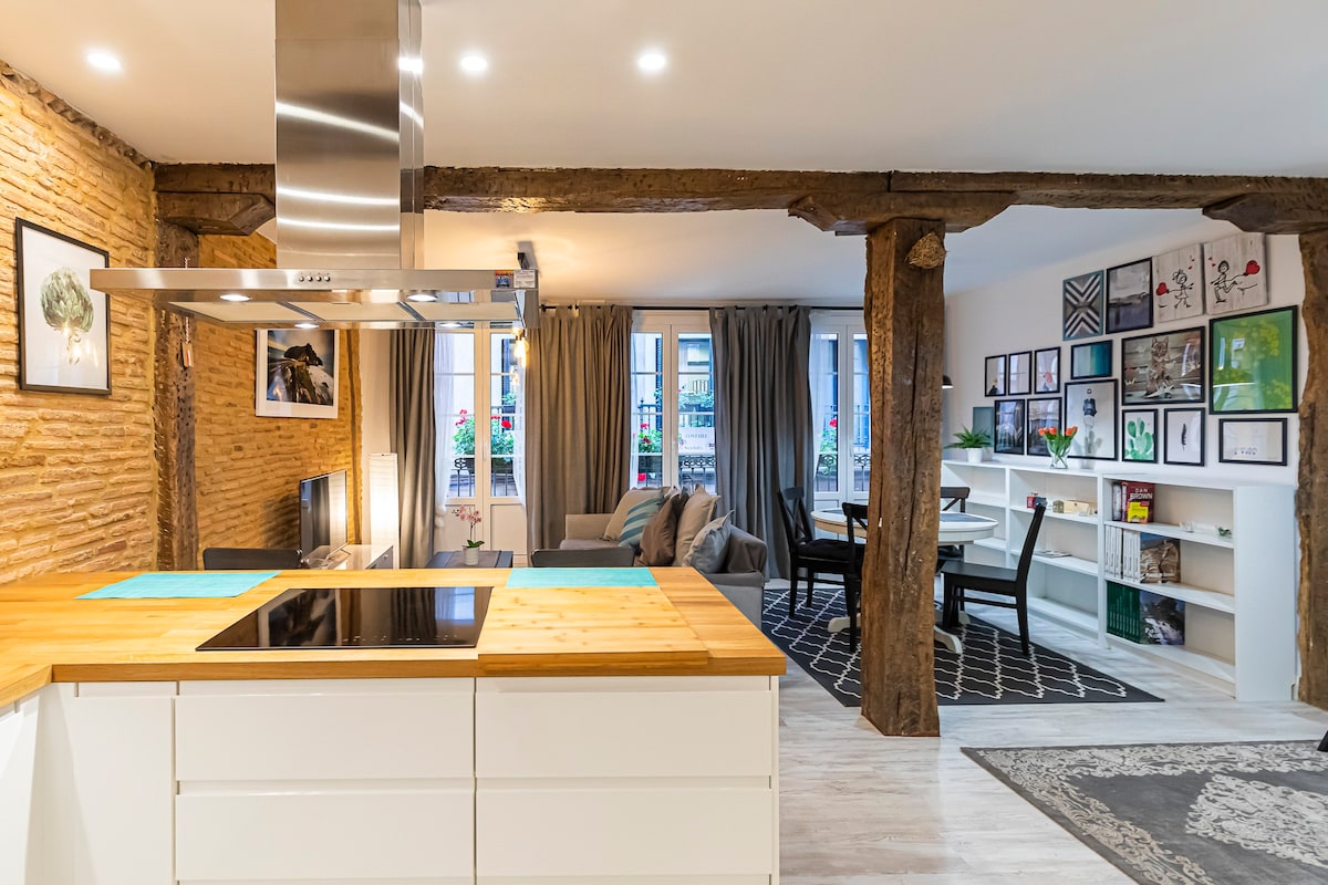 A spacious living area features wooden beams and exposed brick walls. An open kitchen island with a stovetop overlooks a cozy seating arrangement. Sunlight enters through large windows adorned with plants, illuminating a dining table set beside a gallery of framed artwork on the adjacent wall.
