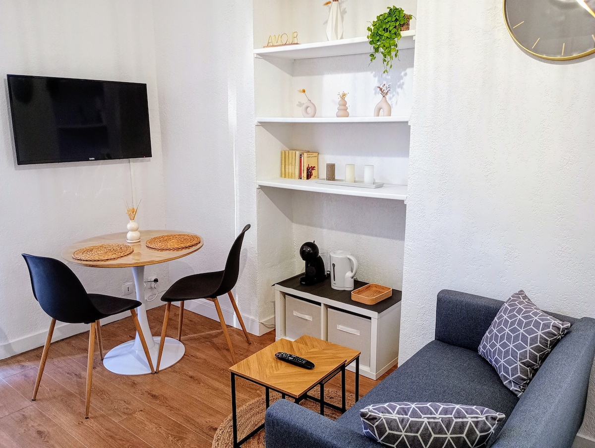 A modern living area is presented with a flat-screen television mounted on the wall. A small round dining table with two black chairs is positioned nearby. A cozy sofa with decorative cushions and a minimalist coffee table are visible, along with a shelf displaying various decorative items.