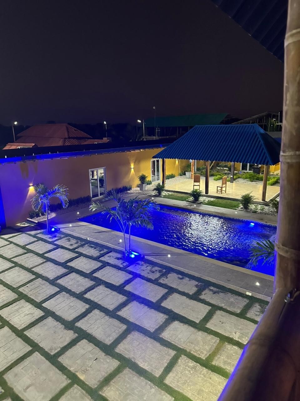 The outdoor area features a well-lit pool surrounded by lush greenery. A stone pathway leads to a covered seating area with a blue roof, while palm trees provide a tropical touch. Soft blue lighting creates a serene ambiance in the evening.