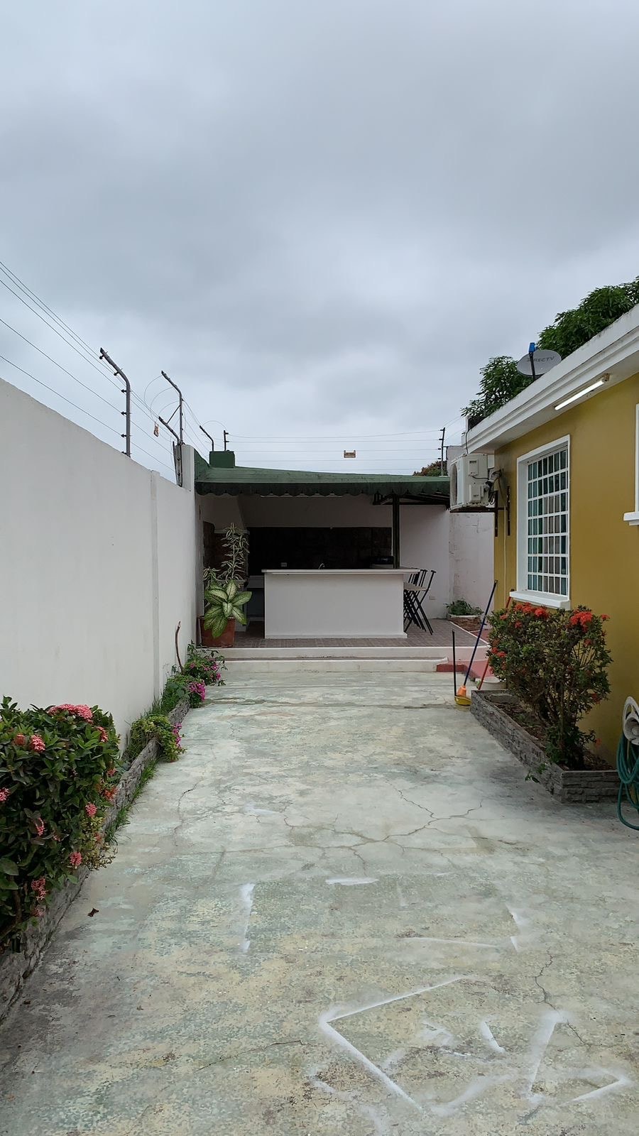 A narrow pathway flanked by low shrubs and flowering plants leads towards an outdoor seating area. A covered bar with high chairs is visible in the background, under a green awning. Gray skies offer a muted backdrop to the serene outdoor space.