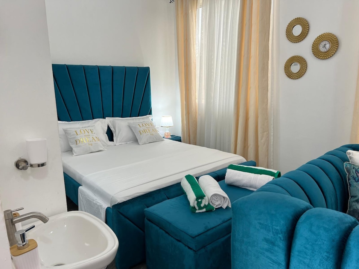 A modern bedroom is highlighted by a plush blue headboard and matching seating. A spacious bed is dressed with crisp linens and decorative pillows. Soft lighting from a bedside lamp complements the light filtering through sheer curtains, while complementary towels are neatly arranged nearby.