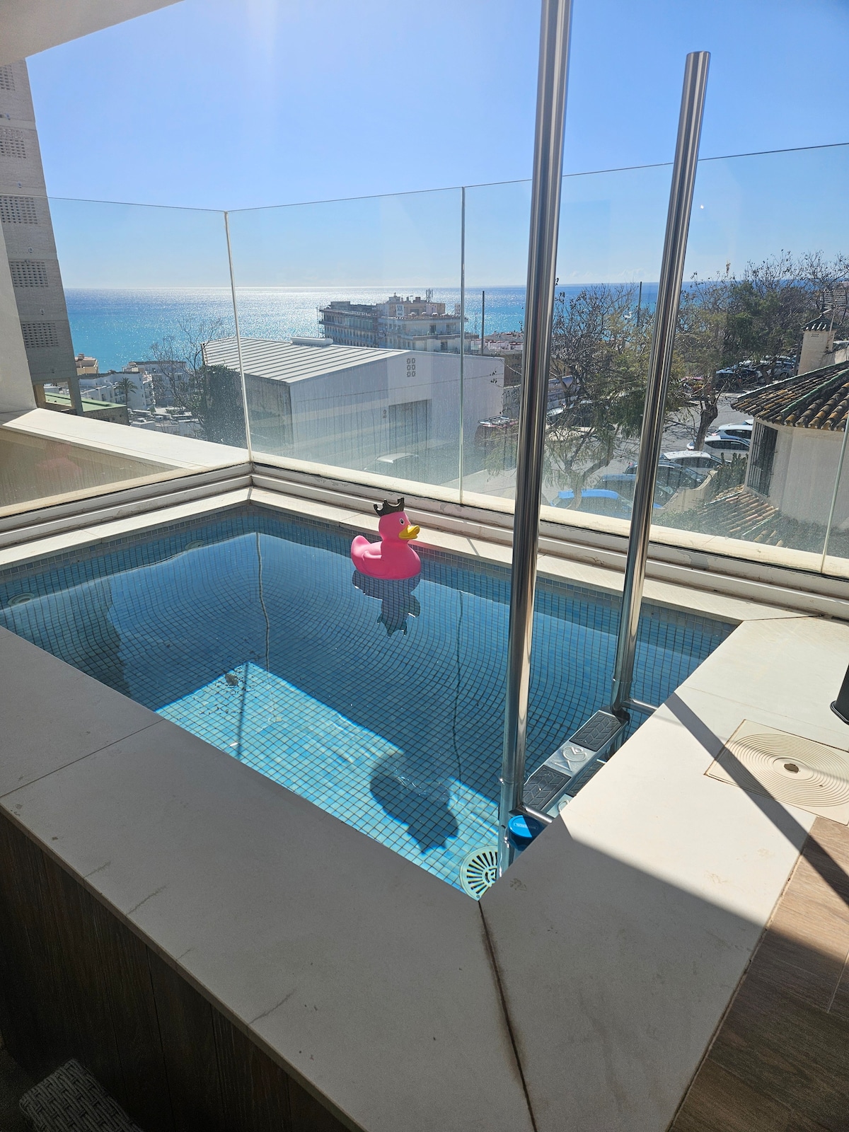 A private terrace features a small pool with clear water, surrounded by glass railings. A colorful inflatable float rests in the water, with a view of the ocean and shoreline visible in the background. The vibrant sky adds to the overall ambiance.