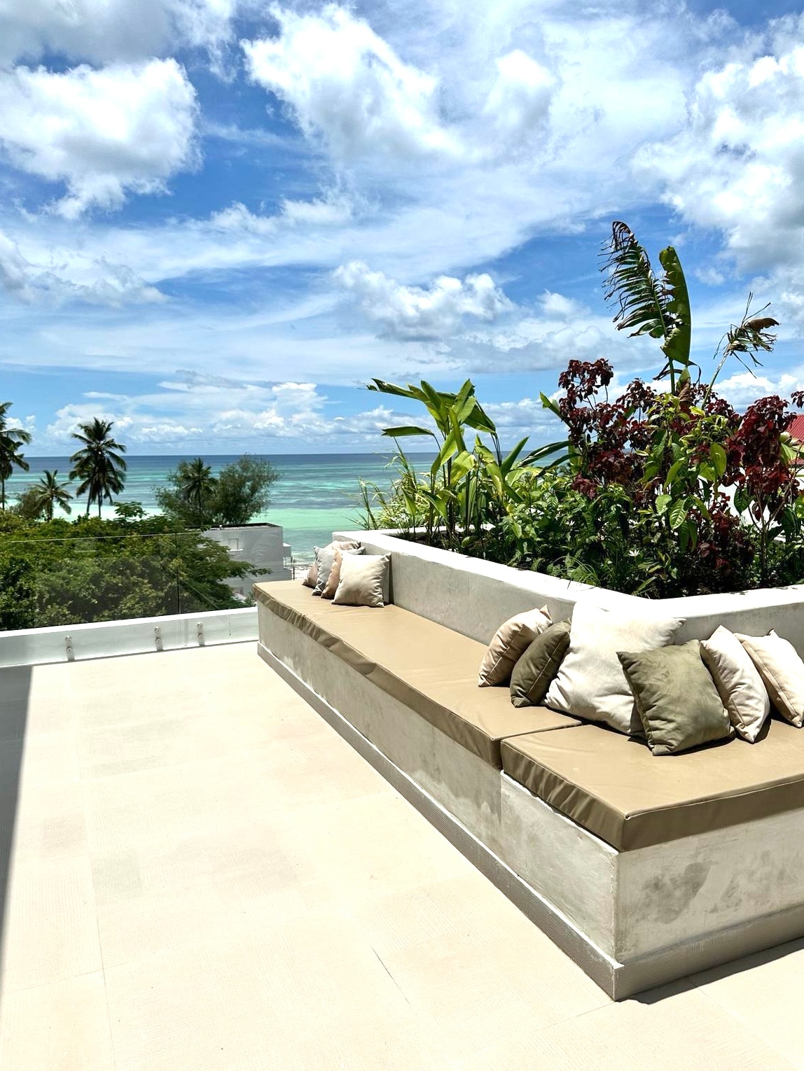 An outdoor lounge area features a spacious, cushioned bench adorned with decorative pillows, surrounded by vibrant tropical plants. The stunning view of the turquoise Indian Ocean is visible in the background, under a bright blue sky dotted with fluffy clouds.