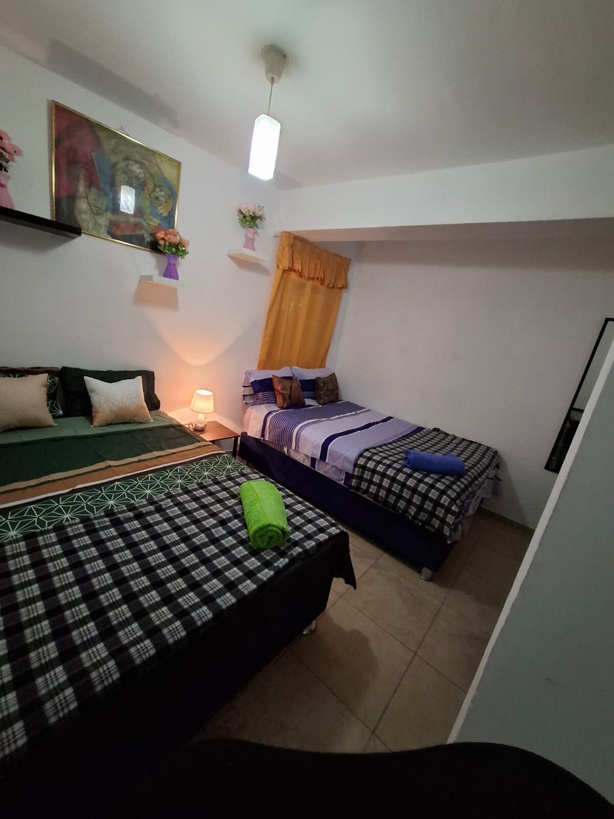 The sleeping area features two beds, one larger and one smaller, both dressed in varied patterned linens. A lamp provides soft lighting on a bedside table, and a vibrant artwork is displayed on the wall above. Natural light filters through a window with sheer curtains.