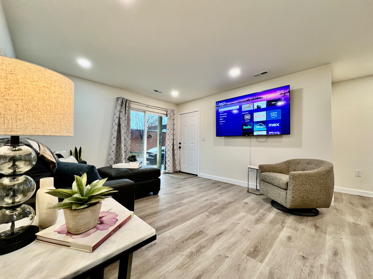 A light-filled living area features a large smart TV mounted on the wall. A comfortable sectional couch and a cozy accent chair are arranged to promote relaxation. Decorative items, including a lamp and a small plant, add a touch of warmth to the modern space.