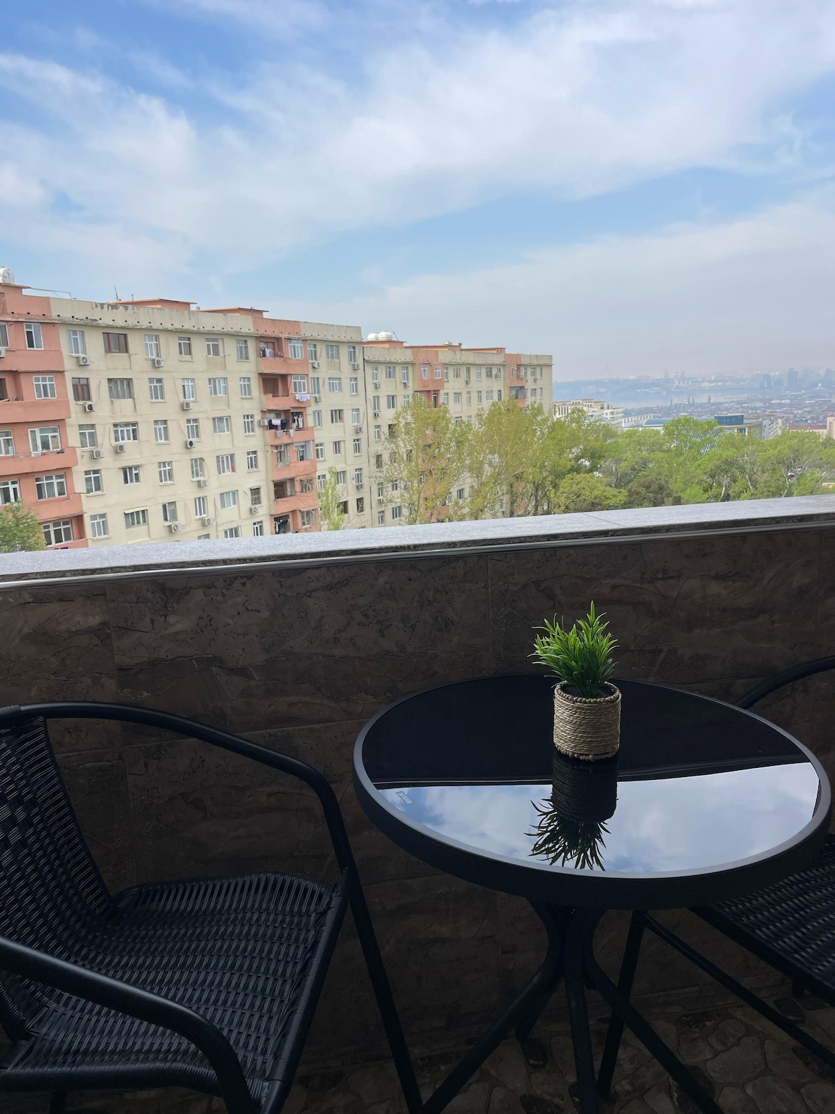 A small balcony area is shown, featuring a round black table and two black wicker chairs. A potted plant is centered on the table. In the background, a view of the city and buildings is visible under a partly cloudy sky.