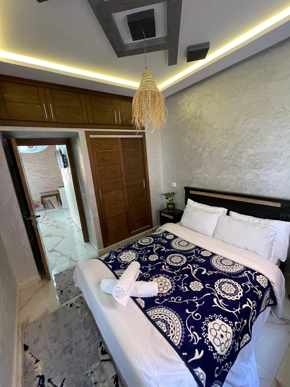 A well-lit bedroom features a spacious bed adorned with a patterned blue and white blanket. Fresh towels are neatly arranged on the bed. White walls and modern light fixtures enhance the room's bright ambiance, while a closet offers ample storage space.
