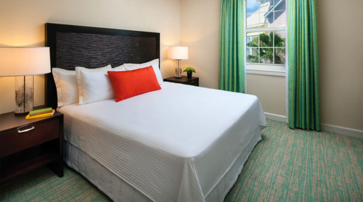 A spacious bedroom is presented with a king-size bed featuring crisp white linens and an accent pillow. Two bedside lamps provide soft lighting. Floor-to-ceiling windows dressed in vibrant green curtains allow natural light, complementing the room's neutral walls and patterned carpeting.