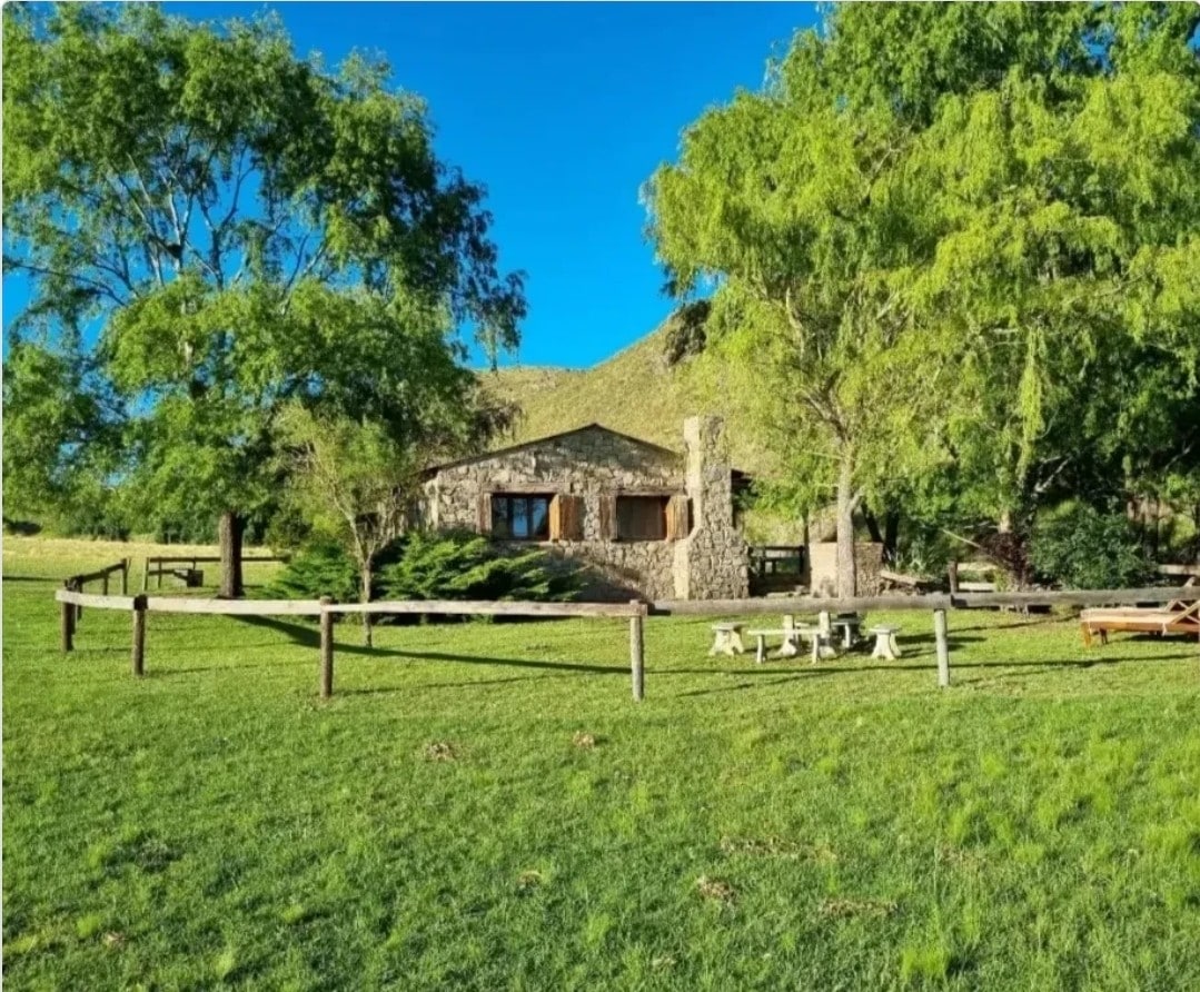 A charming stone cabin is situated amidst lush greenery, framed by tall trees. The cabin features a prominent chimney and multiple windows, allowing plenty of natural light. A flat grassy area in the foreground invites outdoor activities, complemented by rustic seating arranged nearby.