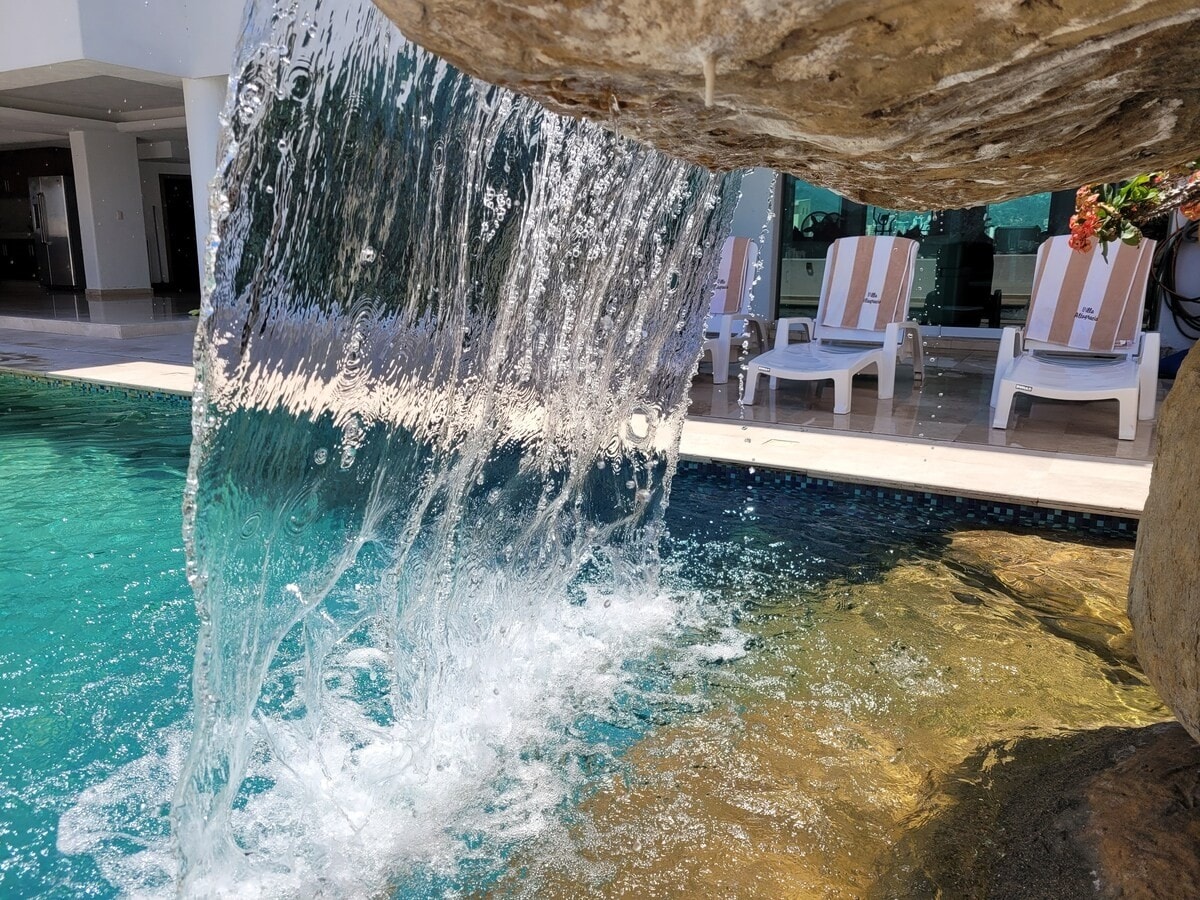 A waterfall cascades over a stone formation, with clear water flowing into a swimming pool. Lounging chairs are positioned nearby, providing a space for relaxation. The pool's surface reflects the sunlight, creating a serene ambiance.