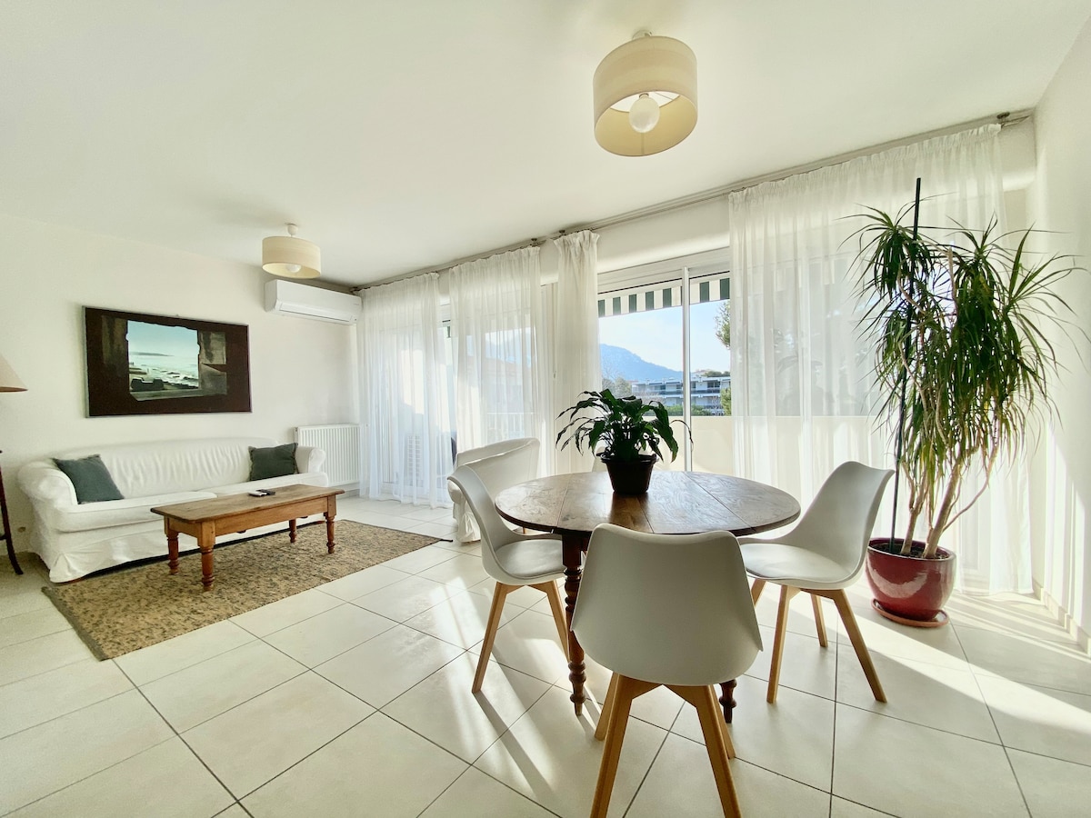 A bright living area features a comfortable white sofa and a wooden coffee table. Natural light filters through sheer curtains, illuminating the space. A round dining table is surrounded by modern chairs, and a green potted plant adds a touch of nature to the room.
