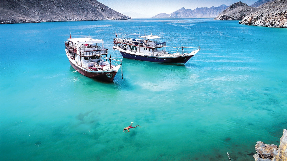 Two boats are anchored in clear turquoise waters, showcasing a vibrant underwater scene. A person is seen swimming nearby, with mountains rising in the background against a clear sky.