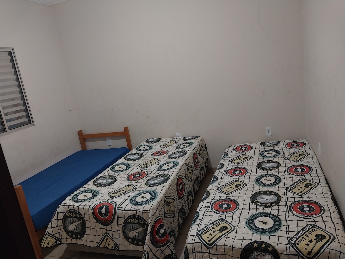 A simple sleeping area featuring two beds covered with patterned bedspreads. One bed is a spacious single with a blue blanket, while the other bed is a single with a monogrammed design. Natural light enters through a small window decorated with horizontal blinds.