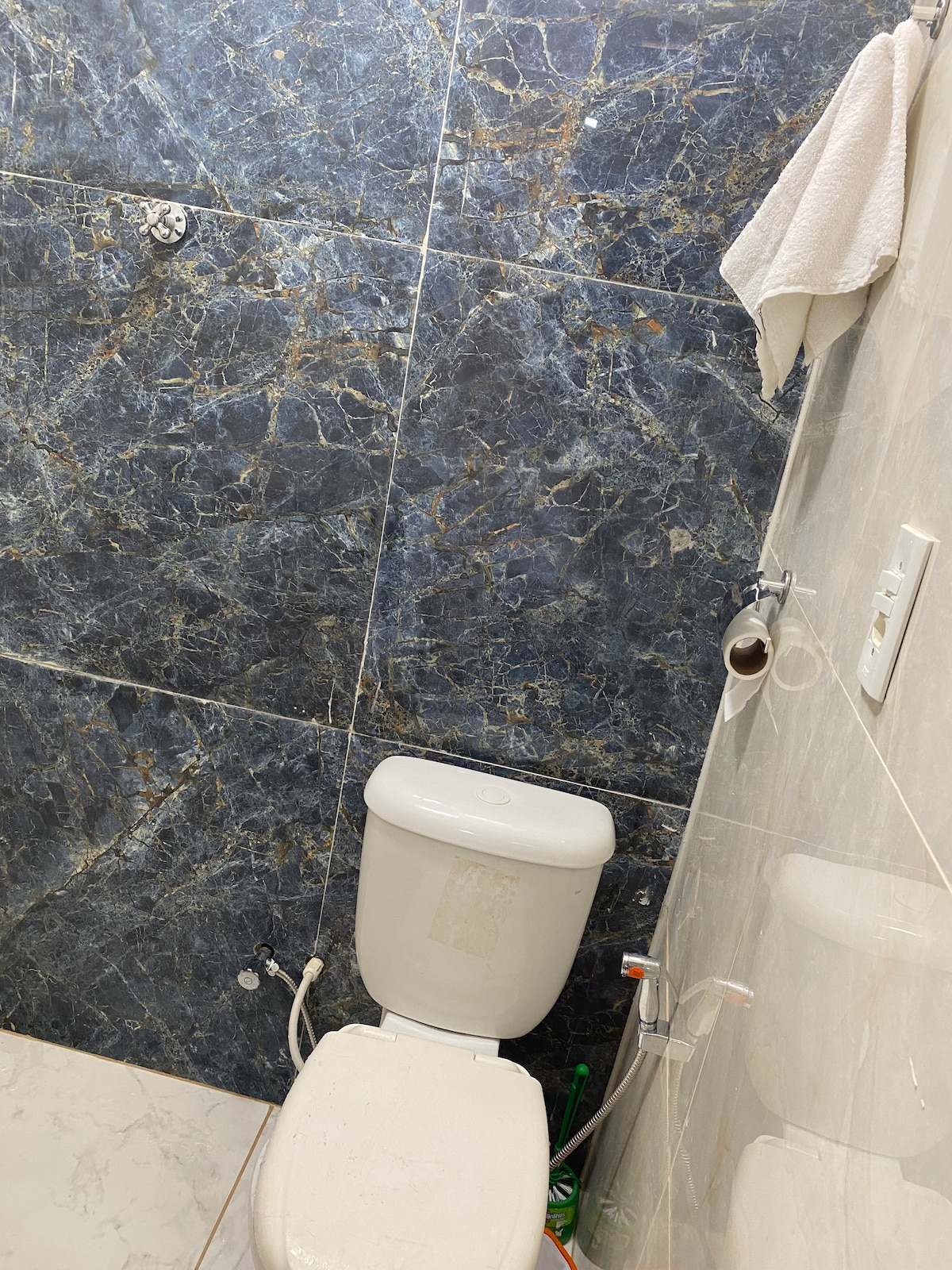 A small bathroom is depicted with a white toilet against a textured blue marble wall. A towel hanging near the top adds functionality, while plumbing fixtures are visible. The floor features light-colored tiles, contributing to a clean and simple aesthetic.
