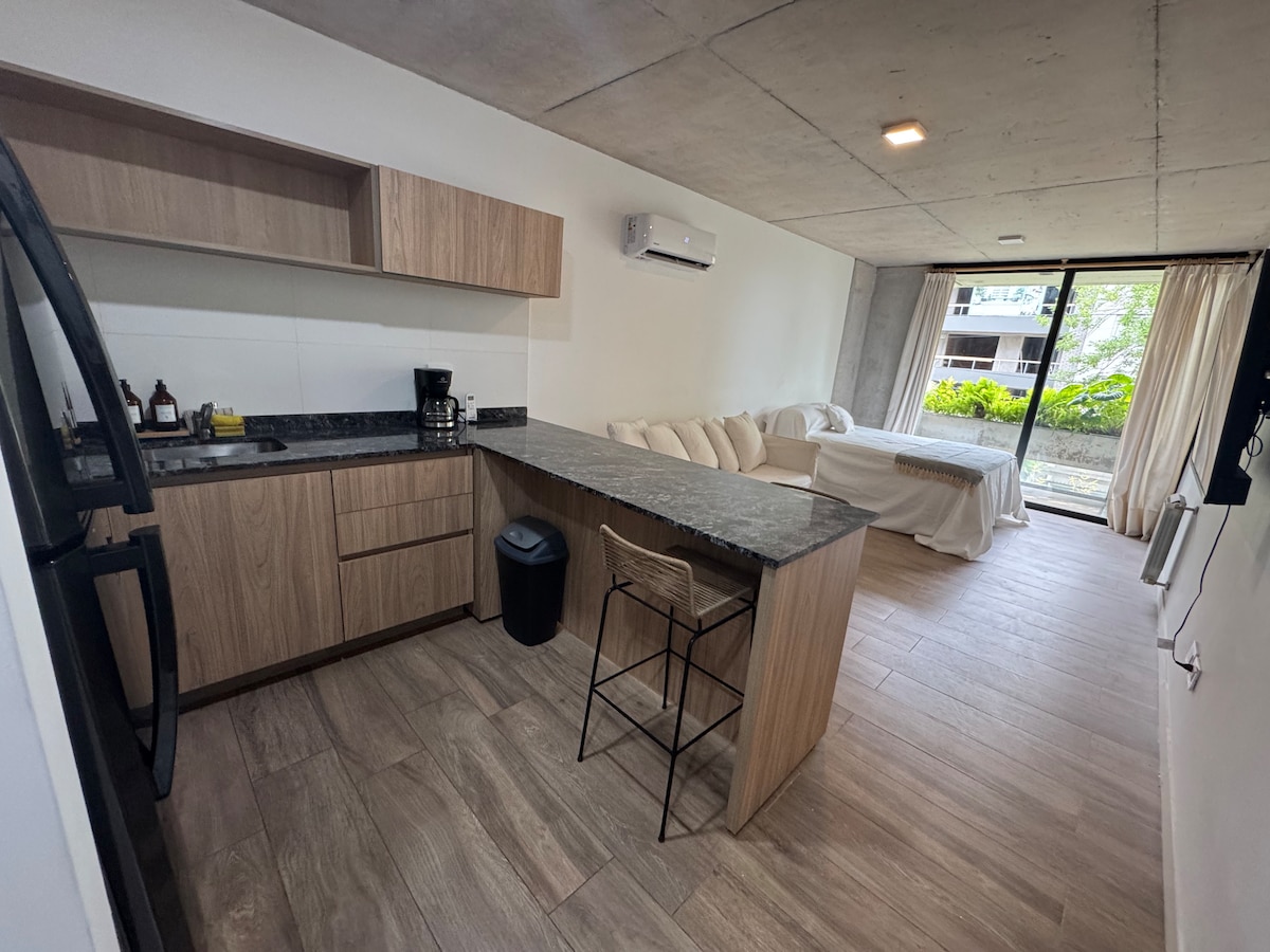 The image showcases a modern, open-concept living space. A kitchen with natural wood cabinetry is visible, along with a dark stone countertop and a bar stool. The area leads to a cozy seating arrangement and large glass doors, allowing natural light to illuminate the space.