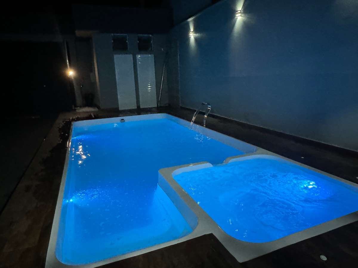 A heated pool is illuminated in soft blue light, featuring steps and a separate area for hydro. Light reflections can be seen on the water's surface, creating a calming atmosphere in a space designed for relaxation.
